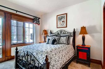 A cozy bedroom featuring a black and white floral bed, a wrought iron bed frame, a rustic nightstand with a red accent, and large windows with wooden blinds.