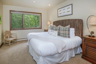 A cozy bedroom in Pines Townhome 10 with a king-sized bed, wicker headboard, and large window overlooking lush green trees.