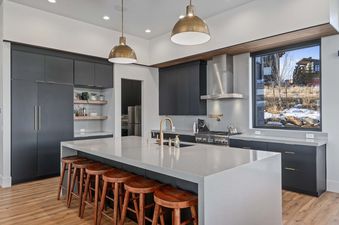 A kitchen with a large island and bar stools, black cabinets, stainless steel appliances, and a view of a snowy mountainside out the window.