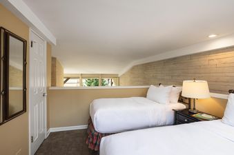 A cozy bedroom with two beds, a nightstand with a lamp, and a wood-paneled accent wall, with a partial view of trees outside.