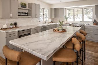 A modern kitchen with grey cabinetry, white marble countertops, and a large island with brown leather bar stools, featuring views of a snowy landscape.