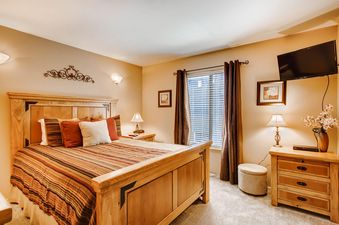 The primary bedroom at Powder Pointe 201A features a large wooden bed, striped bedding, a wall-mounted TV, a nightstand with a lamp, and a window with curtains.