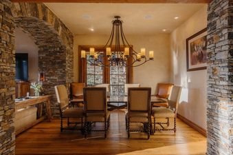 A rustic dining room with a wooden table, upholstered chairs, a wrought iron chandelier, and stone accent walls.