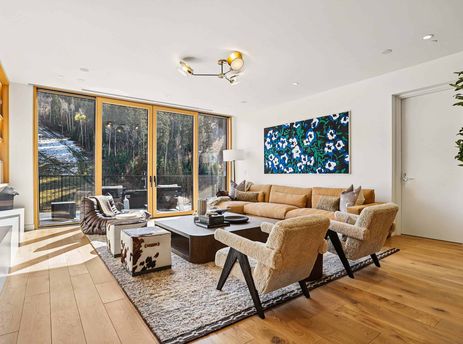 A modern living room with a large brown sectional sofa, sheepskin armchairs, and a view of snow-covered trees through floor-to-ceiling windows.