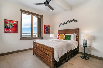 A cozy bedroom with a wooden bed frame, white bedding, and colorful accent pillows, featuring a large window with mountain views and rustic animal artwork on the walls.