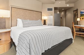 The image shows the Steamboat Grand Deluxe King, a spacious and inviting bedroom with a king-size bed, a bedside table with a lamp, and a desk area.