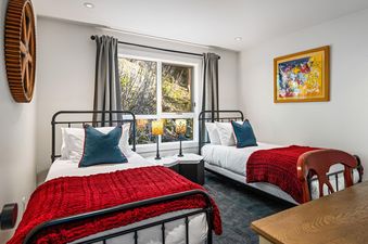 A twin bedroom at St Moritz Townhomes featuring two metal-frame beds with red blankets, a central window with greenery views, a colorful abstract painting, and a unique wooden cog-wheel decor.
