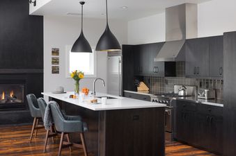 A modern kitchen with dark cabinetry, stainless steel appliances, a white-topped island with bar stools, and a fireplace with a lit fire.