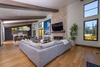 An expansive great room in the Cloud 9 Chalet featuring a light gray sectional sofa, a stone fireplace with a mounted TV, and large windows showcasing an autumnal landscape.