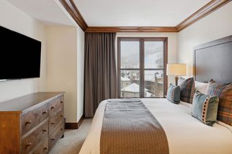 A cozy bedroom with a large bed, a wooden dresser, and a window offering a view of snowy mountains and buildings.