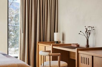 A modern bedroom in the Mollie Aspen Park View Corner Suite features a comfortable bed, a stylish wooden desk with a chair, and a large window showcasing a snowy mountain view.