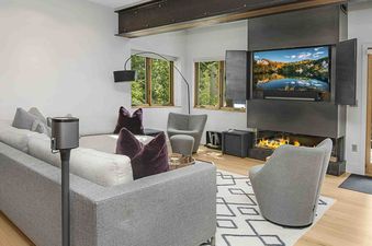 The living room of 1175 A Casolar Del Norte, featuring a fireplace, a large flat screen TV, and comfortable furniture, including a large couch and two chairs. The room has large windows that provide ample natural light and views of the outdoors.