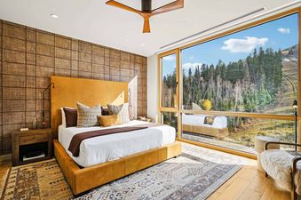A modern bedroom at Founders Place 2502 features a yellow upholstered bed, textured accent wall, and large windows with a scenic mountain view.