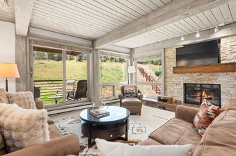 A cozy living room at Enclave 211 with a stone fireplace, large windows overlooking a green hillside, and comfortable seating.