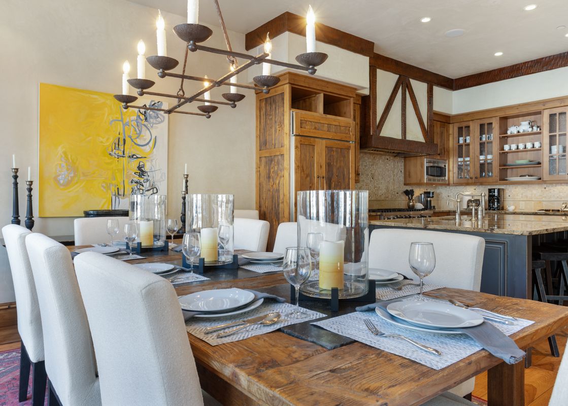 A rustic dining table set with white chairs and a chandelier,  with a kitchen in the background.