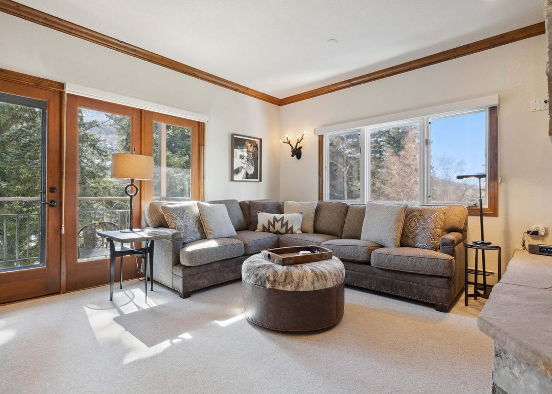 A cozy living room featuring a large sectional sofa, a round cowhide ottoman with a tray, and a wall of windows and glass doors opening to a balcony with mountain views.