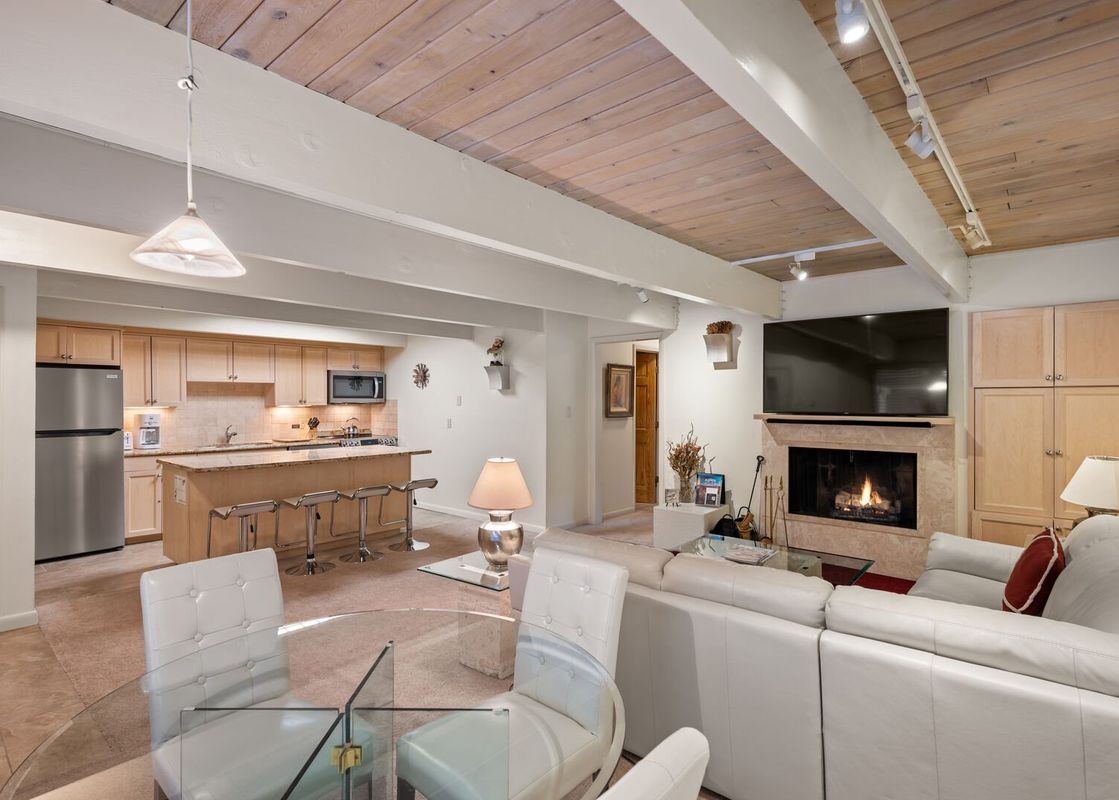 An open-plan living space with a kitchen, dining area, and lounge, featuring a stone fireplace and a wooden beamed ceiling.