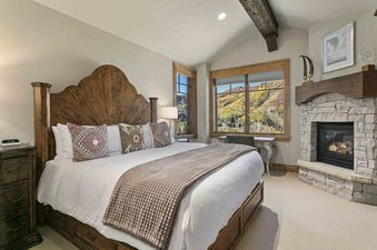 The primary bedroom at Silver Strike Lodge 703 features a wooden king-sized bed with decorative pillows and a textured throw, a stone fireplace with a wooden mantel, and large windows offering stunning views of autumn mountains.
