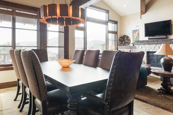 A formal dining room with a large dark wood table, eight upholstered chairs, and a striking circular chandelier, set against a backdrop of large windows offering mountain views.