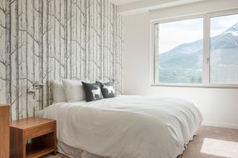 A modern bedroom with a bed featuring white bedding and throw pillows with animal silhouettes, set against a birch tree wallpaper, and a large window overlooking a mountain landscape.