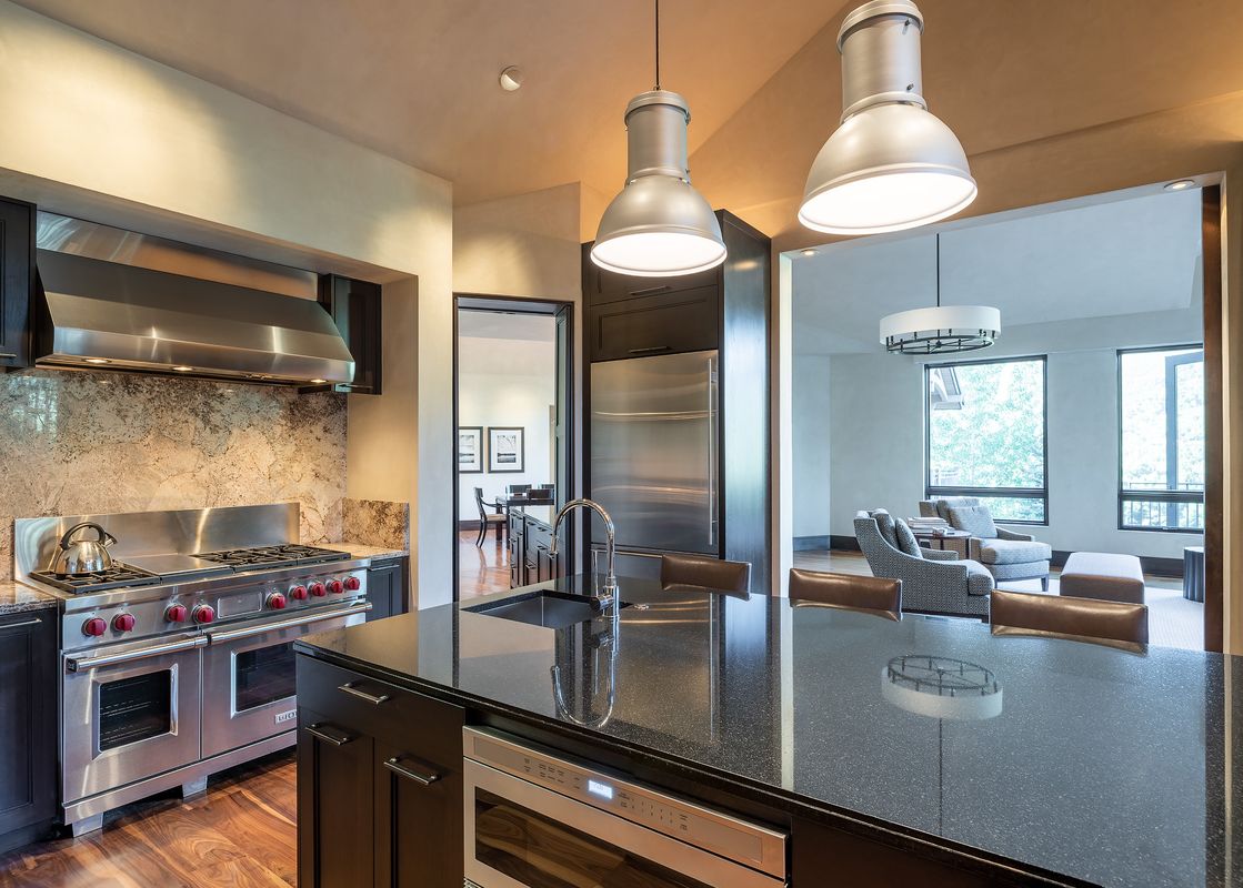 A modern kitchen with dark cabinetry, black granite countertops, and stainless steel appliances flows into a bright living room.