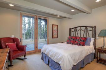 Moonlight Basin Lodging 38A Cowboy Heaven features a comfortable bedroom with a king-size bed and a rustic wood headboard. The room also features a cozy reading chair, and a large window with views of the trees outside.