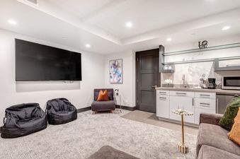 A modern living room with a large TV, a cozy chair, and a large sectional couch. There is also a small kitchen and a small bar area.