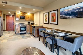 A cozy kitchen and dining area in Copperbottom 205 with wooden cabinets, stainless steel appliances, a dining table set for four, and a wall-mounted TV showing a winter mountain scene.