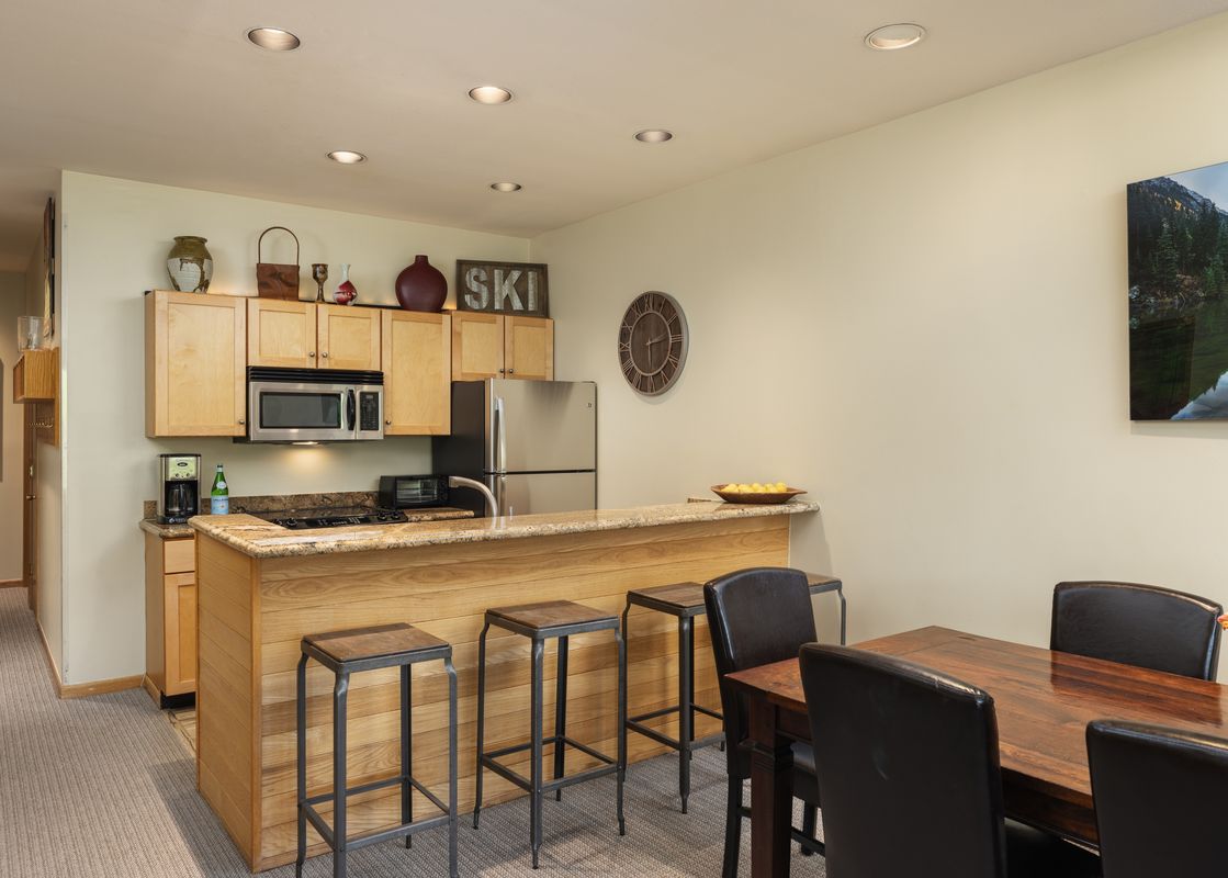 The kitchen and dining area at Villas at Snowmass Club 1515, featuring light wooden cabinets, stainless steel appliances, a breakfast bar, and a dining table with dark leather chairs.
