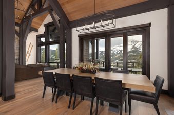 A spacious dining room at Summit View Chalet Big Sky with a large wooden table, dark chairs, and expansive windows offering a stunning view of snow-capped mountains.