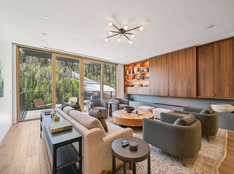 A modern living room at Founders Place 2405 with a beige sofa, grey armchairs, and a round wooden coffee table, featuring a view of a snowy forest through large glass doors.