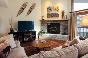 A cozy living room at the Ranch at Steamboat RA520 with a stone fireplace, comfortable sofas, and a flat-screen TV, featuring rustic decor and a view of a snowy landscape through a large window.