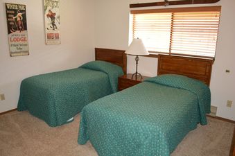 The twin bedroom features two single beds with teal bedspreads and wooden headboards, a bedside table with a lamp, and a large window with blinds.