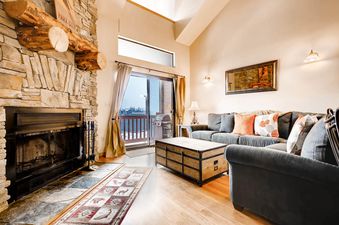 A cozy living room at Powder Pointe 303B with a stone fireplace, gray sofas, a trunk coffee table, and a balcony view of a snowy outdoor scene.