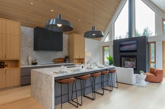 A modern kitchen with a large island and bar seating, a fireplace, and a large window with a view of the forest