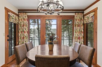 A dining room with an oval wooden table and brown leather chairs, featuring large windows overlooking a snow-covered lake and pine trees.