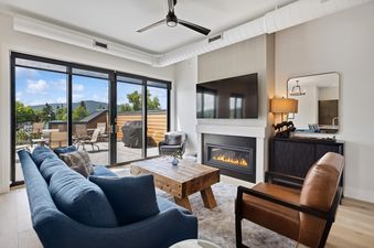 A modern living room with a blue sofa, wooden coffee table, fireplace, and flat-screen TV, opening to an outdoor patio with mountain views.