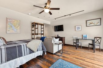 An inviting open-plan living and sleeping area in Cana Loft 203, featuring a bed, sofa, television, and dining table, all under the warm glow of a ceiling fan.