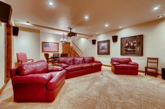 The Bear Tracks Lodge living area features three large red leather sofas, a gaming table with chairs, and wall art, all under recessed lighting and a ceiling-mounted projector.