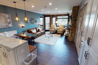 A living room with a leather couch, two armchairs, a coffee table, and a fireplace, with an adjacent kitchen with granite countertops, white cabinets, and a wooden table.