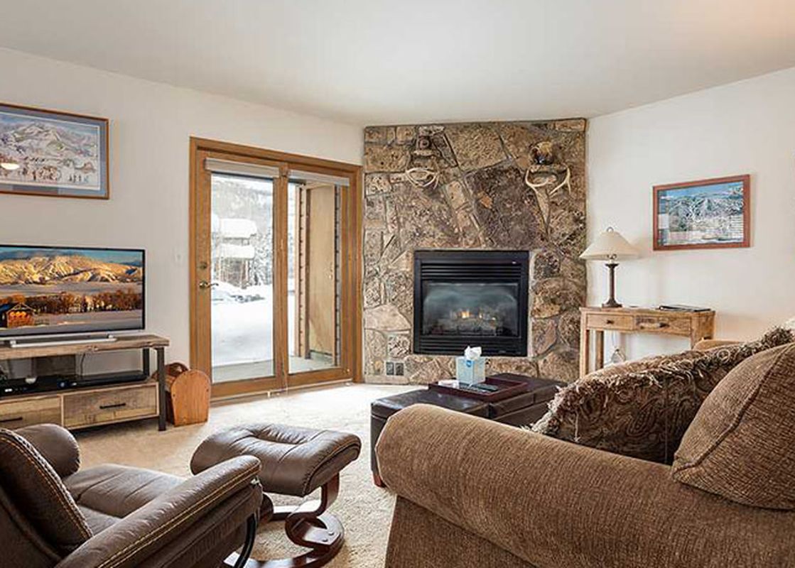 A cozy living room in Pines Condominiums Two Bedroom with a stone fireplace, comfortable seating, and a flat-screen TV, leading to a snowy patio.