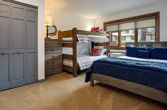 A cozy bedroom at Ranch at Steamboat RA215 features a queen bed with blue bedding, a wooden bunk bed with plaid and red accents, a chest of drawers, and a window with blinds overlooking other buildings.