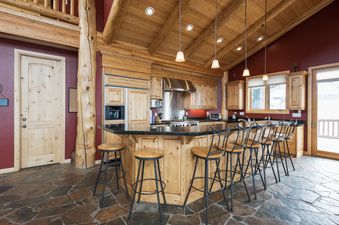 A rustic kitchen with natural wood cabinetry, a dark granite island with bar stools, stainless steel appliances, a vaulted wooden ceiling, and flagstone flooring.