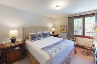 The bedroom at Manor Vail 280 features a comfortable king-size bed with a cozy blanket, two bedside tables with lamps, and a large window offering a beautiful view of the surrounding trees.