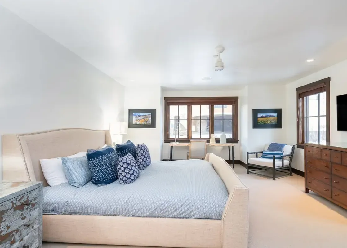 A large bedroom with a king-sized bed with a cream headboard, a wooden dresser with drawers, a flat screen TV, a window with a view, and a chair in the corner of the room.