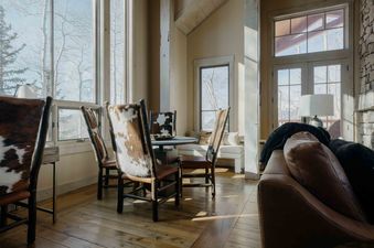 A dining area with a round table and chairs with cowhide upholstery, overlooking a snowy landscape through large windows.