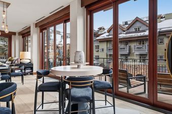 The dining area at Solaris 2A South features a round table surrounded by dark blue chairs, set against a backdrop of large windows with snowy mountain and neighborhood views.