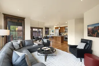 A wide shot of the modern open-concept living area and kitchen at On Trails Edge, featuring a large gray sectional sofa, a black marble coffee table, and a well-appointed kitchen with wooden cabinetry and stainless steel appliances.