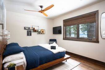 A cozy bedroom at West Shore Hideaway with a wooden bed frame, blue and white bedding, a ceiling fan, and a large window overlooking a lake.