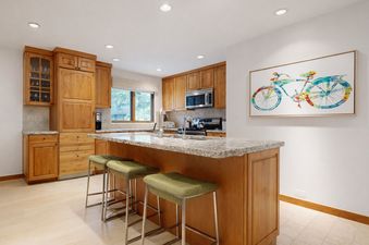 A modern kitchen with natural wood cabinets, a granite island with barstools, and colorful bicycle artwork on the wall.